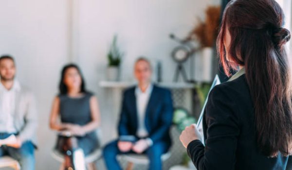 Businesswoman holding a clipboard and calling out candidates for a job interview. Shot of manager standing opposite group of business people sitting in line, waiting for a job interview meeting, reading their names and calling them out.